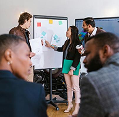 Three people present on a whiteboard during a meeting while others listen in the foreground.