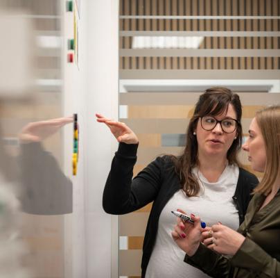 Two women work together in a meeting, one writing notes on a whiteboard as they discuss ideas.