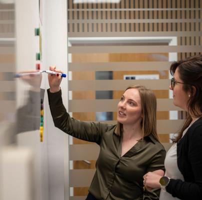 Two women collaborate in a meeting, one writing ideas on a whiteboard while the other observes and contributes.