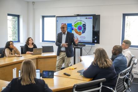 A man stands at the front of a room pitching to a diverse group of people, who are seated and listening.