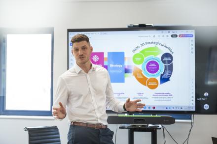 A man stands at the front of a room pitching to a diverse group of people