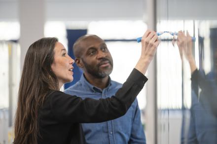 A woman writes on a glass board in an office setting while a man stands beside her, engaged in the discussion.