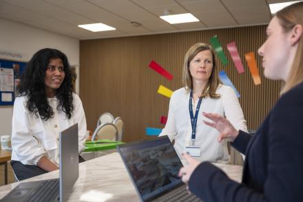Three women sit together in a meeting, engaged in discussion and sharing ideas, with a laptops on the table in front of them.