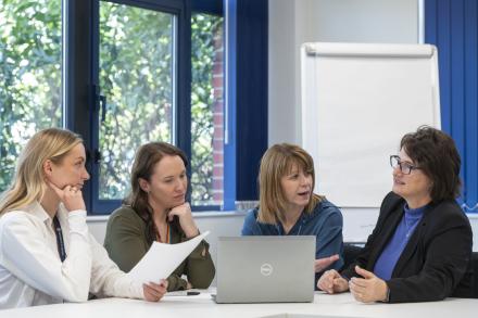 Four women sit together in a meeting, discussing ideas with an open laptop on the table.