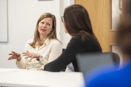 Two women sit together in an open-plan office, engaged in a discussion.