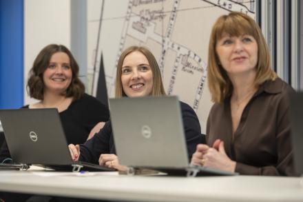Three women sit side by side in an office space, listening to a discussion with open laptops on the table.