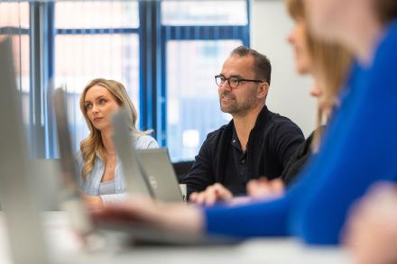 A man listens attentively to colleagues speaking in an open-plan office discussion.