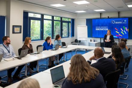 A woman delivers a pitch in an office while a diverse group of men and women seated around tables listen attentively.