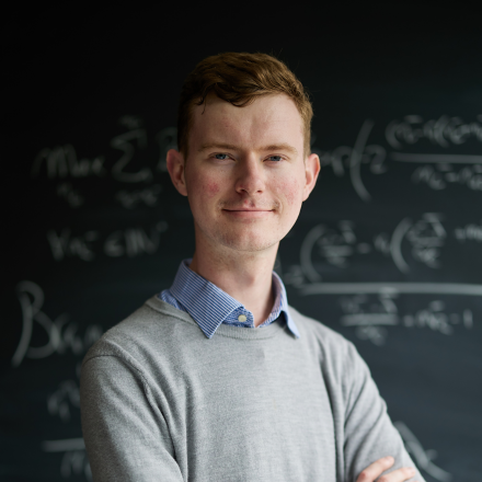 A man crossing his arms smiling facing the camera. He wears a grey jumper over a blue shirt and is stood in front of a chalkboard background with scientific equations written on it.
