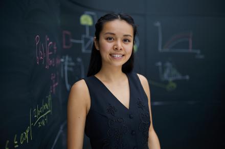 A woman facing the camera smiling with dark hair pulled back, wearing a black v-neck top. She stands in front of a dark chalkboard background with scientific equations written on it.