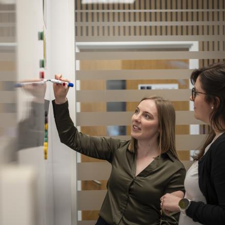 Two women collaborate in a meeting, with one writing on a whiteboard while the other looks on.
